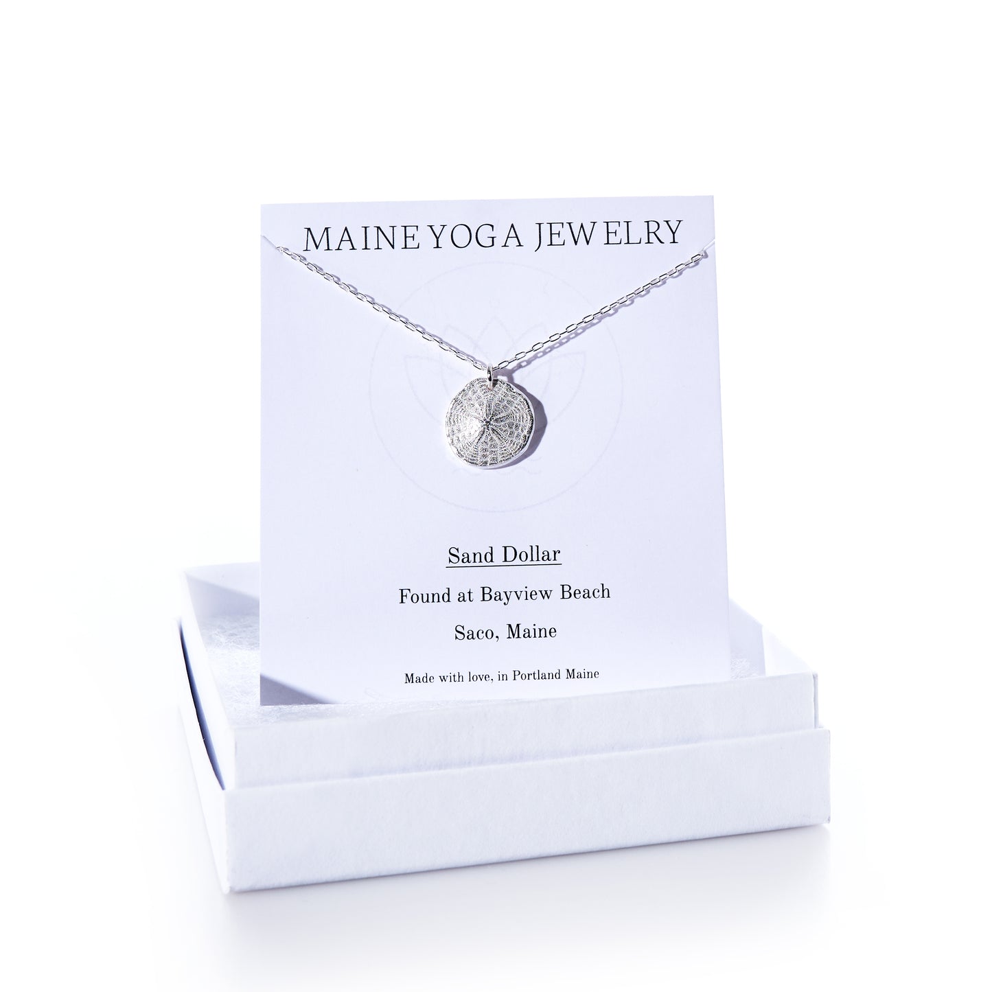 A Pendant Necklace - Sand Dollar Silver by Maine Yoga Jewelry rests on a card inside a white box. The card notes that the sand dollar was found at Bayview Beach, Saco, Maine, emphasizing the brands sustainable craftsmanship inspired by Maines natural beauty. by Sea Bags