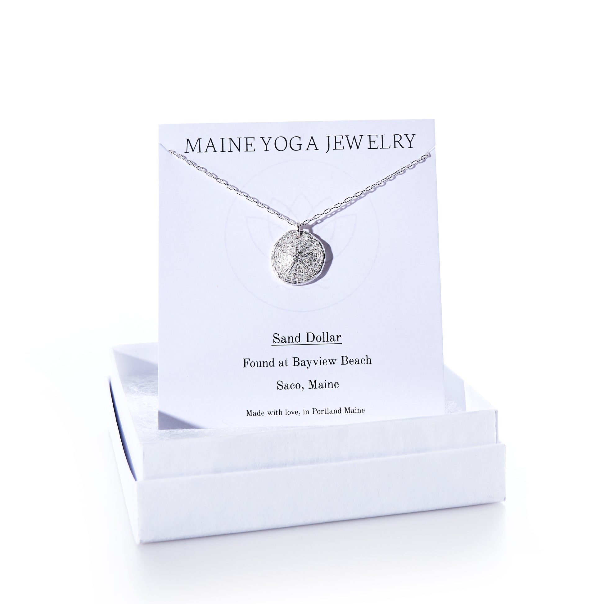 A Pendant Necklace - Sand Dollar Silver by Maine Yoga Jewelry rests on a card inside a white box. The card notes that the sand dollar was found at Bayview Beach, Saco, Maine, emphasizing the brands sustainable craftsmanship inspired by Maines natural beauty. by Sea Bags