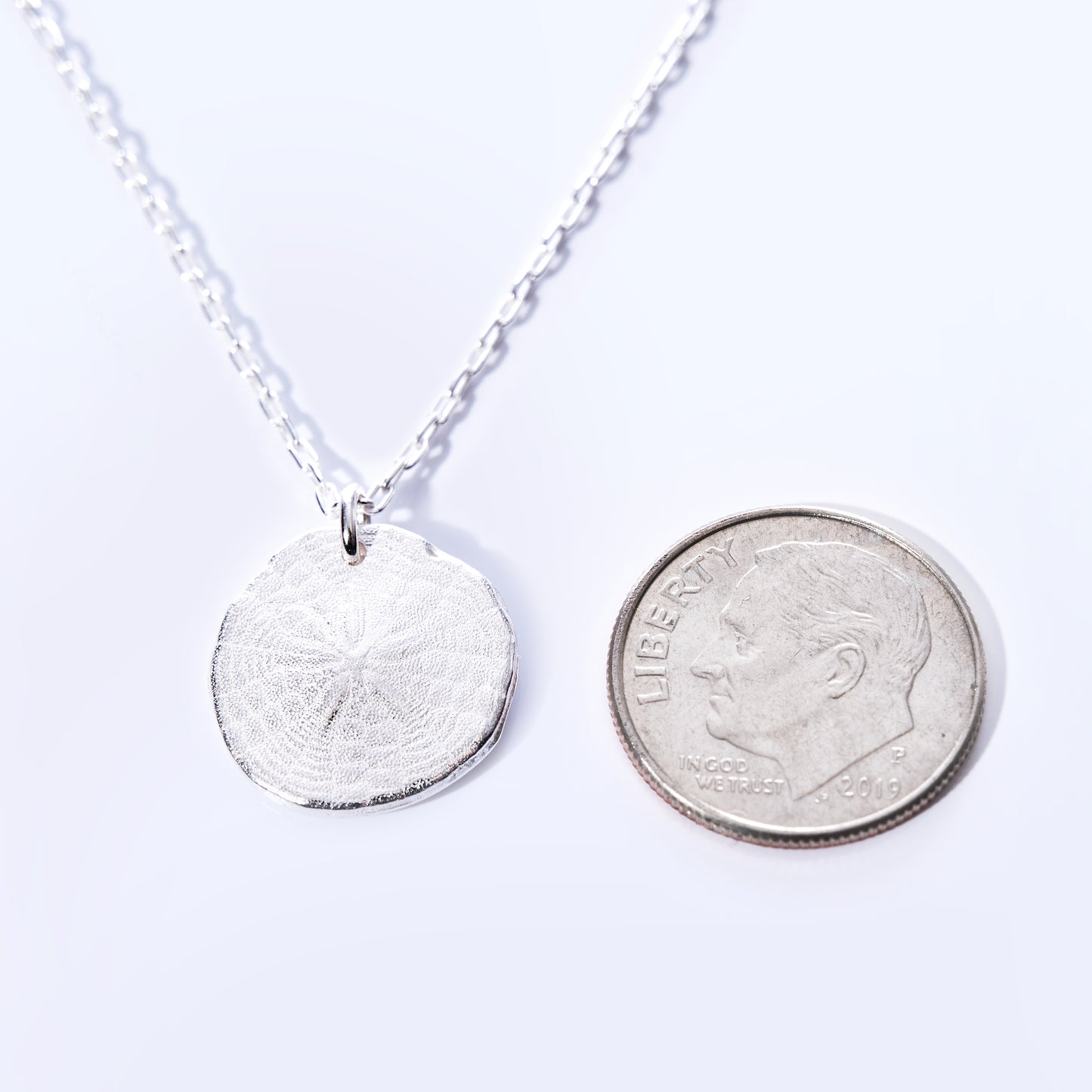 The Pendant Necklace - Sand Dollar Silver by Maine Yoga Jewelry features a round, textured silver sand dollar charm—handcrafted in Maine with sustainable craftsmanship—and is pictured next to a U.S. dime on a white background for size reference. by Sea Bags