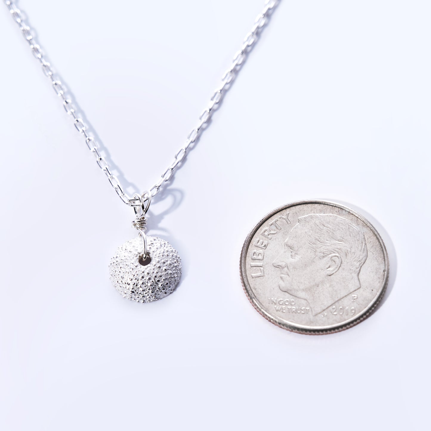 A Pendant Necklace - Tiny Sea Urchin Silver by Maine Yoga Jewelry is displayed beside a US dime on a white background. Handcrafted in Maine, this round, textured pendant captures coastal elegance and artisanal charm as it hangs from a delicate silver chain. by Sea Bags