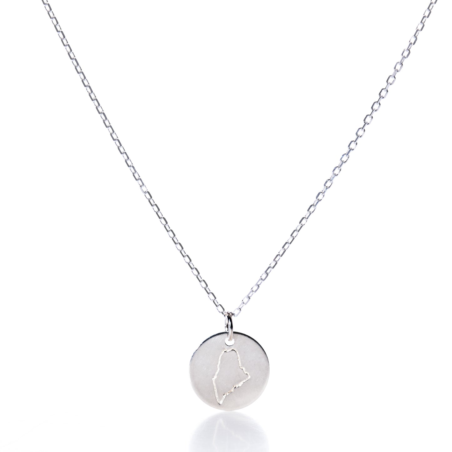 Showcased on a white background, the Pendant Necklace - Silver Maine by Maine Yoga Jewelry features a round silver pendant engraved with an outline of the state of Maine. This elegant necklace beautifully captures the spirit and charm of New England, making it a perfect piece of coastal jewelry. by Sea Bags