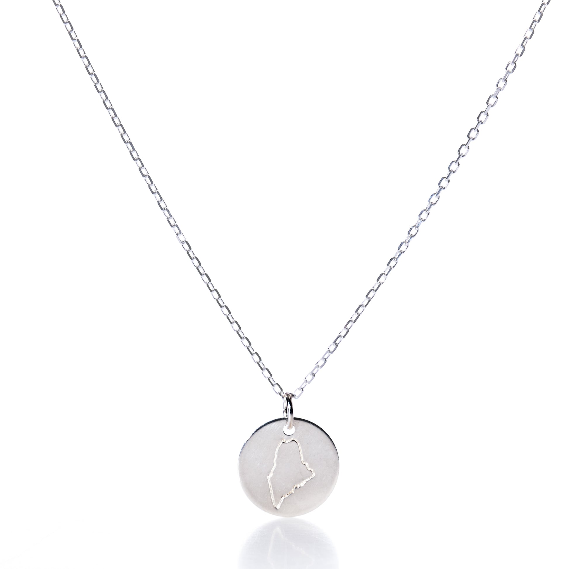 Showcased on a white background, the Pendant Necklace - Silver Maine by Maine Yoga Jewelry features a round silver pendant engraved with an outline of the state of Maine. This elegant necklace beautifully captures the spirit and charm of New England, making it a perfect piece of coastal jewelry. by Sea Bags