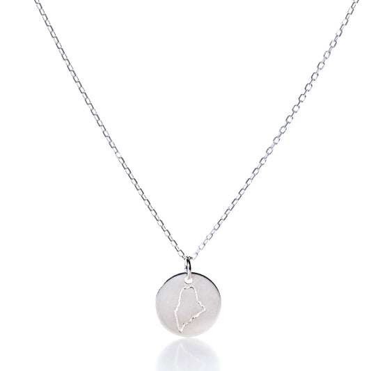 Showcased on a white background, the Pendant Necklace - Silver Maine by Maine Yoga Jewelry features a round silver pendant engraved with an outline of the state of Maine. This elegant necklace beautifully captures the spirit and charm of New England, making it a perfect piece of coastal jewelry. by Sea Bags