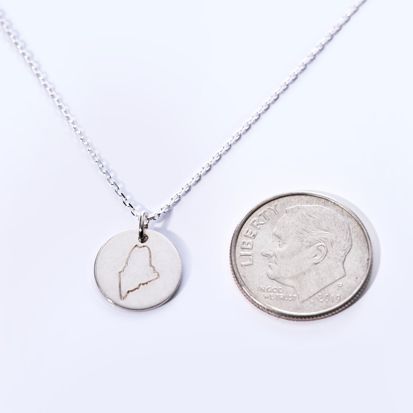 The Pendant Necklace - Silver Maine by Maine Yoga Jewelry features a circular silver charm engraved with an outline of Maine, shown beside a US dime for size comparison—making it an ideal coastal jewelry piece for anyone who loves the state. by Sea Bags
