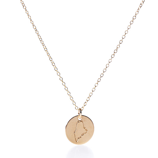 Pendant Necklace - Gold Maine by Maine Yoga Jewelry features a round gold charm engraved with the outline of Maine, artfully displayed on a plain white background. Expertly crafted in Maine, this piece showcases timeless elegance and distinctive craftsmanship. by Sea Bags