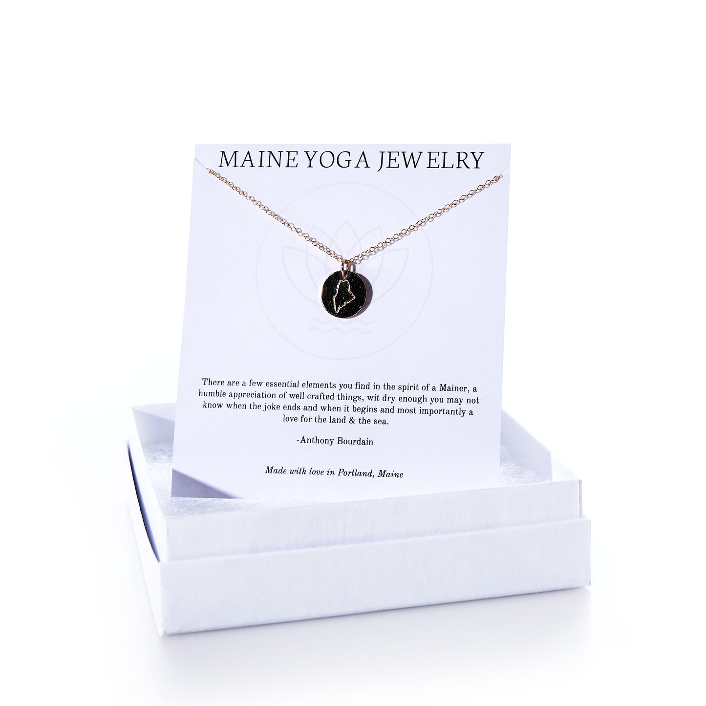 Pendant Necklace - Gold Maine by Maine Yoga Jewelry features a handcrafted round black pendant made from recycled sailcloth, elegantly suspended on a gold necklace. It comes displayed on a white card labeled “Maine Yoga Jewelry,” beautifully presented in a white jewelry box. Thoughtfully made in Maine, this sustainable piece combines eco-friendly materials with sophisticated design. by Sea Bags