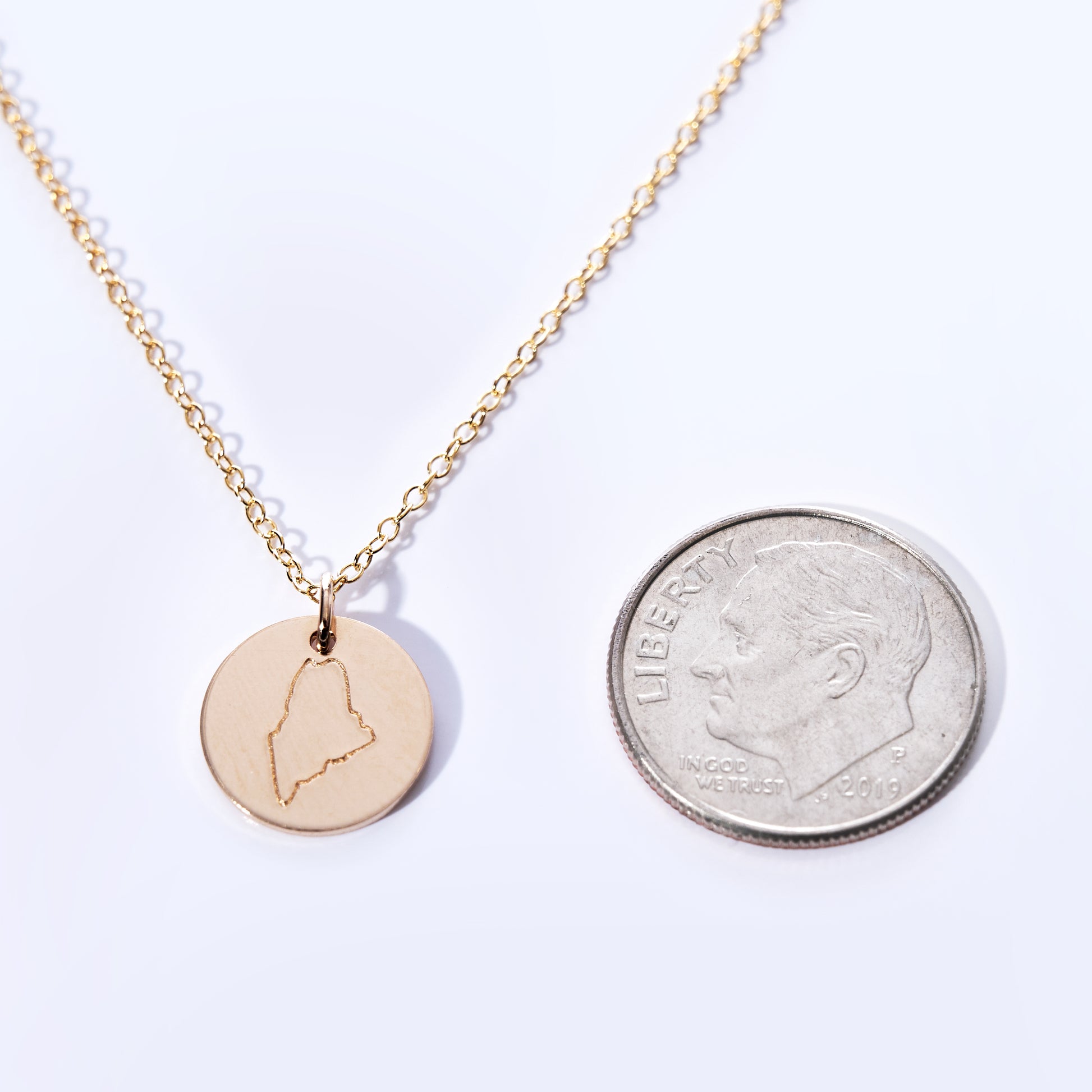 Presenting the Pendant Necklace - Gold Maine by Maine Yoga Jewelry: a refined gold necklace featuring a round pendant engraved with the outline of the state of Maine. Shown beside a US dime for scale on a white background, this elegant piece is carefully crafted in Maine and beautifully embodies both local pride and timeless style. by Sea Bags