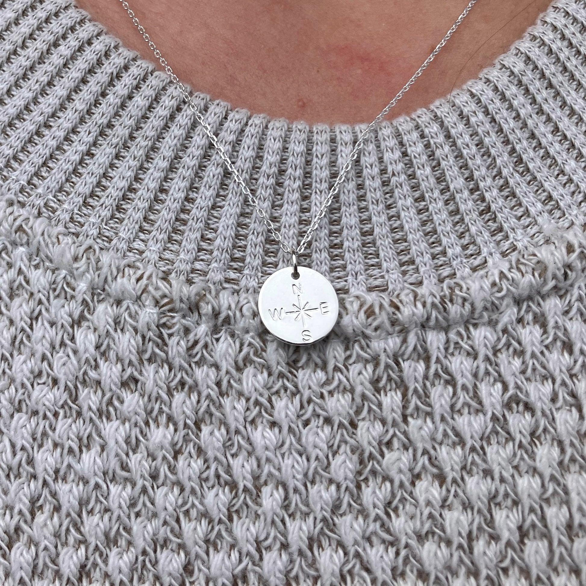 The Pendant Necklace - Silver Compass Rose by Maine Yoga Jewelry features a round silver compass pendant with N, S, E, and W letters, elegantly worn over a light gray knitted sweater—an exquisite nautical necklace beautifully crafted in Maine. by Sea Bags