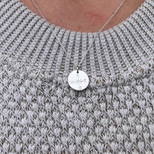 The Pendant Necklace - Silver Compass Rose by Maine Yoga Jewelry features a round silver compass pendant with N, S, E, and W letters, elegantly worn over a light gray knitted sweater—an exquisite nautical necklace beautifully crafted in Maine. by Sea Bags