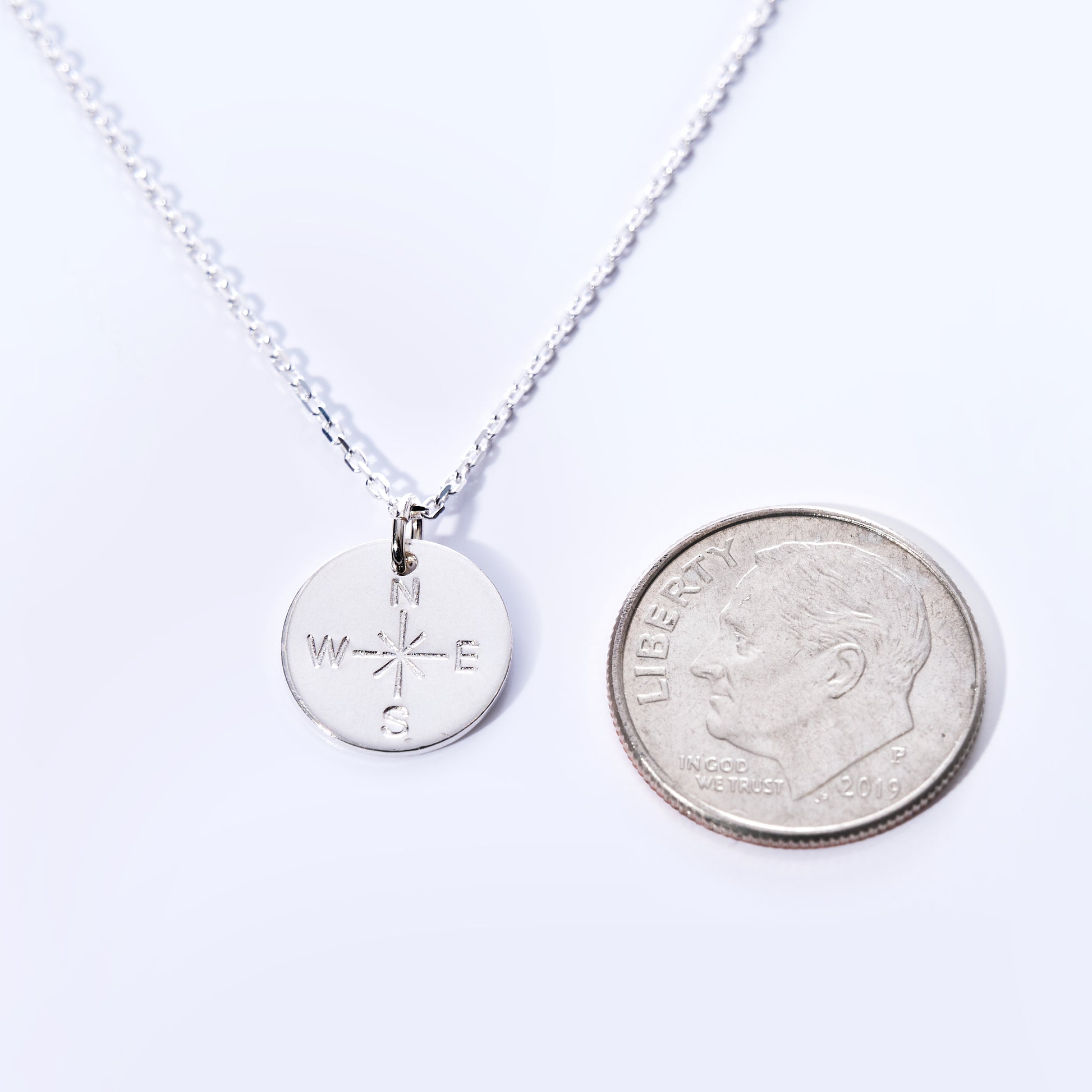 A Pendant Necklace - Silver Compass Rose by Maine Yoga Jewelry is displayed next to a US dime for size comparison on a white background. This nautical necklace features a compass pendant and is thoughtfully crafted and proudly made in Maine. by Sea Bags