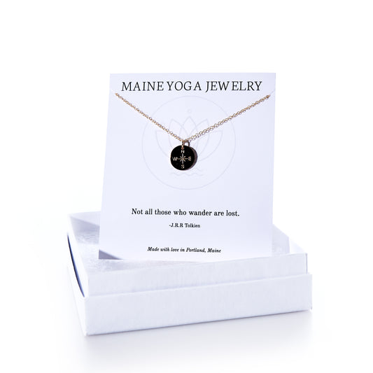 A Pendant Necklace - Gold Compass Rose by Maine Yoga Jewelry is presented on a card featuring a Tolkien quote, with its round black pendant inscribed with WANDER. This nautical necklace comes beautifully packaged in a white box and is crafted in Maine. by Sea Bags