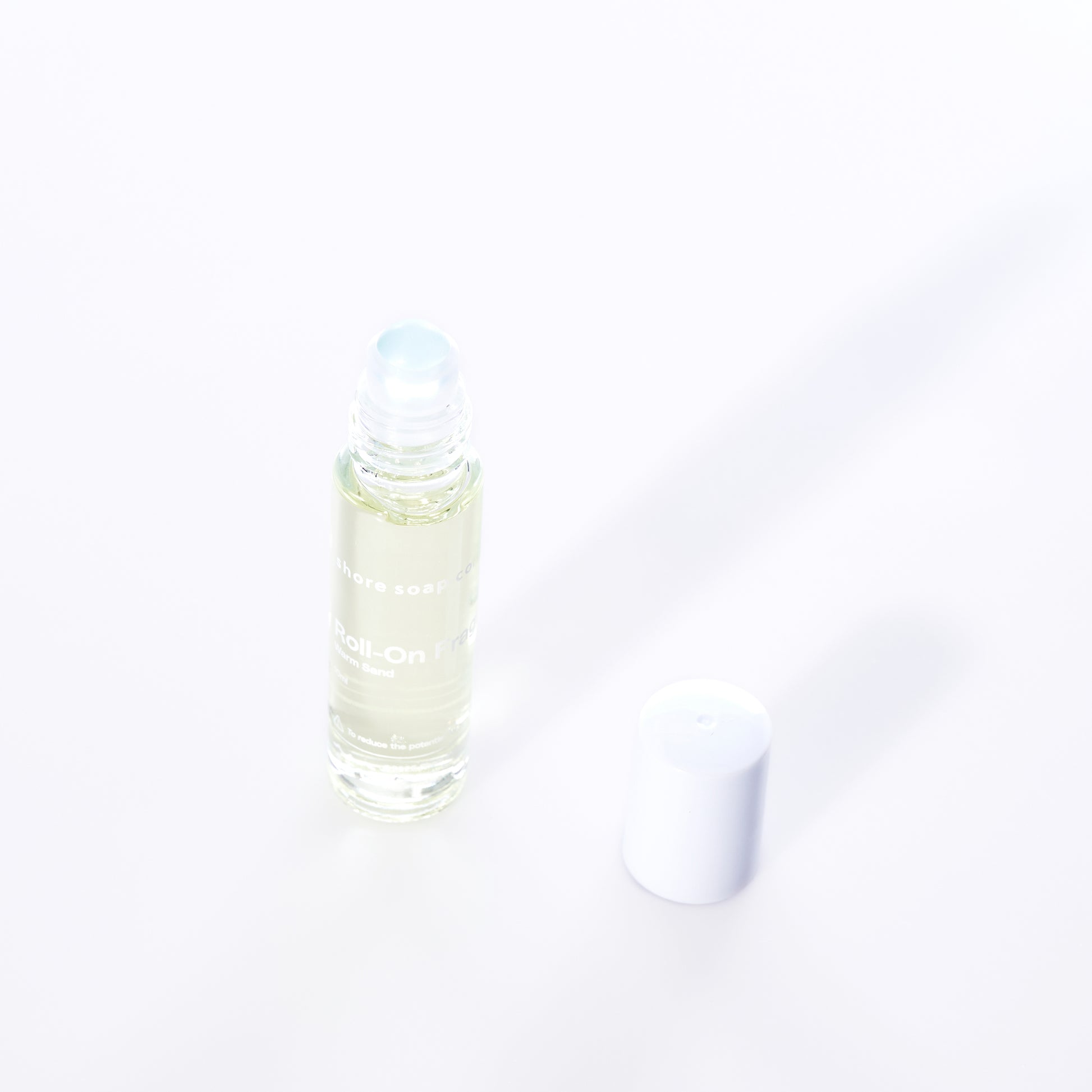 A clear glass roll-on bottle of Shore Soap Cos Warm Sand Eco-Friendly Roll-On Fragrance, featuring an alcohol-free jasmine scent, with a white plastic cap placed beside it on a white background. by Sea Bags