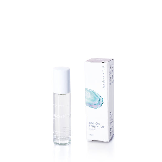 The Atlantic Eco-Friendly Roll-On Fragrance by Shore Soap Co is displayed in a clear glass bottle with a white cap, standing upright beside its matching white packaging box on a white background. This convenient roll-on design makes it an ideal travel perfume, letting you enjoy your favorite scent wherever you go. by Sea Bags