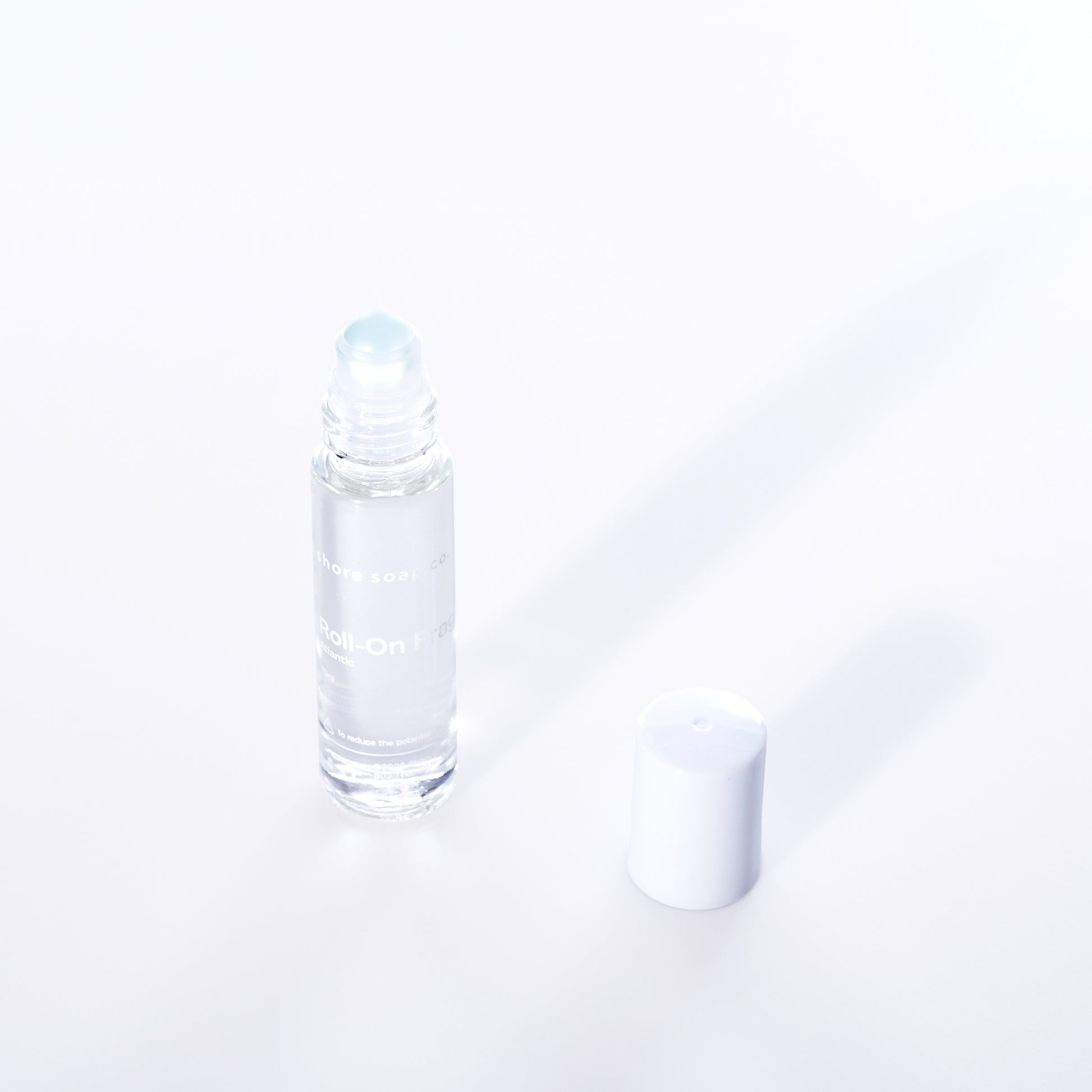 The Atlantic Eco-Friendly Roll-On Fragrance by Shore Soap Co is shown in a clear glass bottle with a white cap placed beside it on a white background, making it perfect for carrying your favorite alcohol-free perfume as a convenient travel fragrance. by Sea Bags