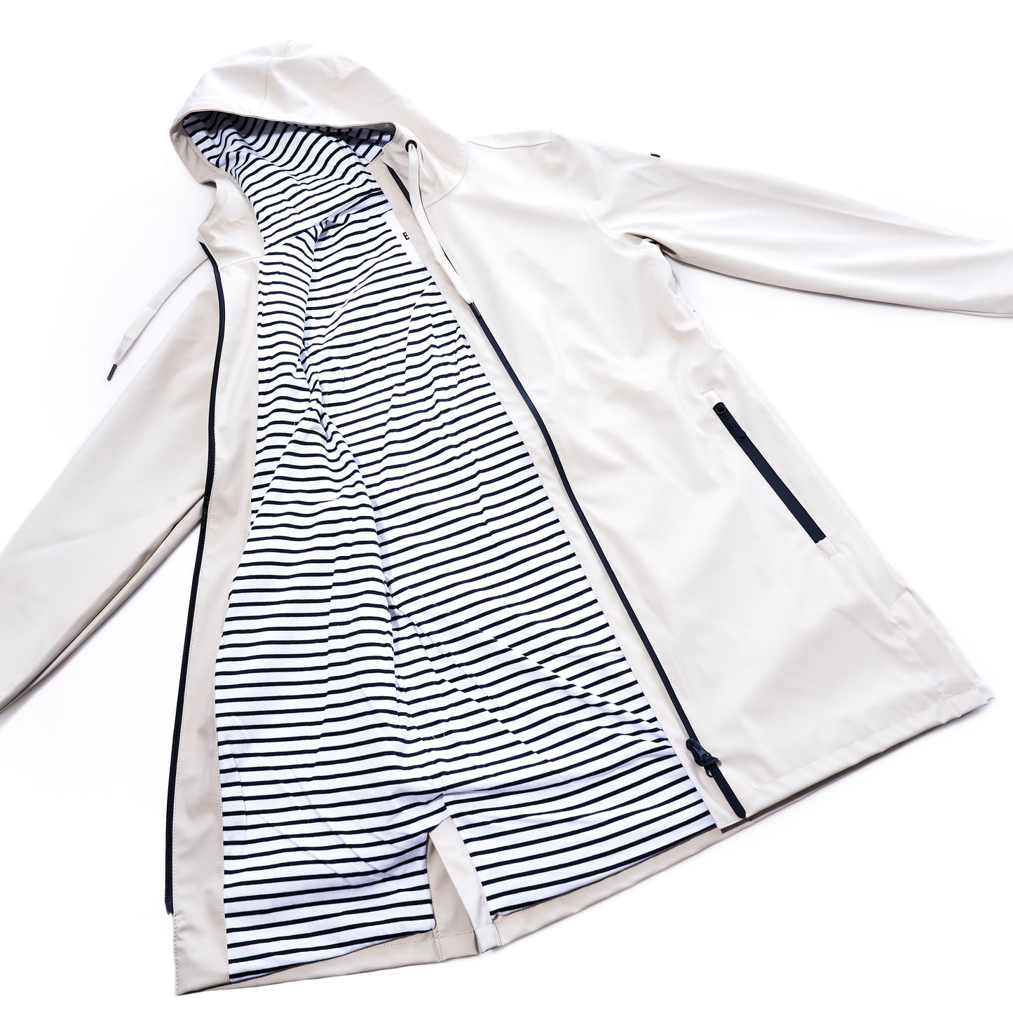 The Harborside Waterproof Raincoat - Moonstruck by true is shown laid flat, unzipped to reveal its black and white striped inner lining and black-trimmed zipper pockets. This beige, unisex raincoat features a waterproof shell, seamlessly blending style with all-weather practicality.