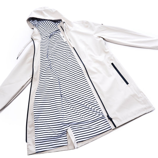 The Harborside Waterproof Raincoat - Moonstruck by true is shown laid flat, unzipped to reveal its black and white striped inner lining and black-trimmed zipper pockets. This beige, unisex raincoat features a waterproof shell, seamlessly blending style with all-weather practicality.
