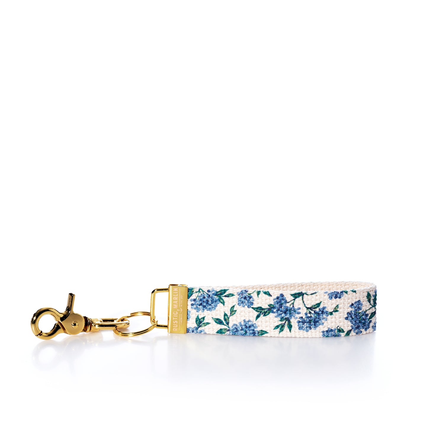 Introducing the Rustic Marlin Keychain - Blueberry by Rustic Marlin: This elegant keychain features gold hardware and a fabric strap adorned with a blue floral pattern on a white background, beautifully displayed on a white surface. Designed to double as a chic bag charm, its sturdy brass key clip keeps your keys secure and within easy reach. by Sea Bags