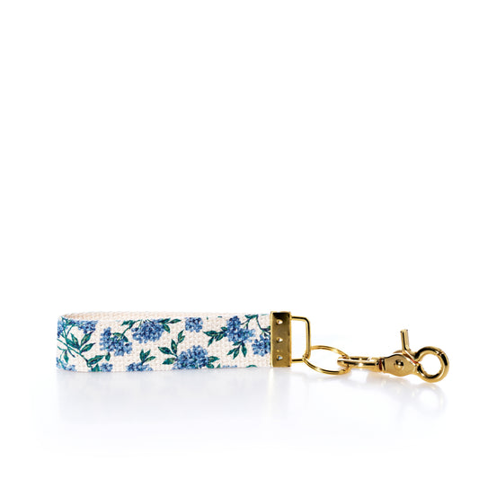 The Rustic Marlin Keychain - Blueberry by Rustic Marlin features a charming blue floral pattern and gold keyring, displayed on a white background—perfect as a stylish bag charm or unique blueberry keychain accessory. by Sea Bags