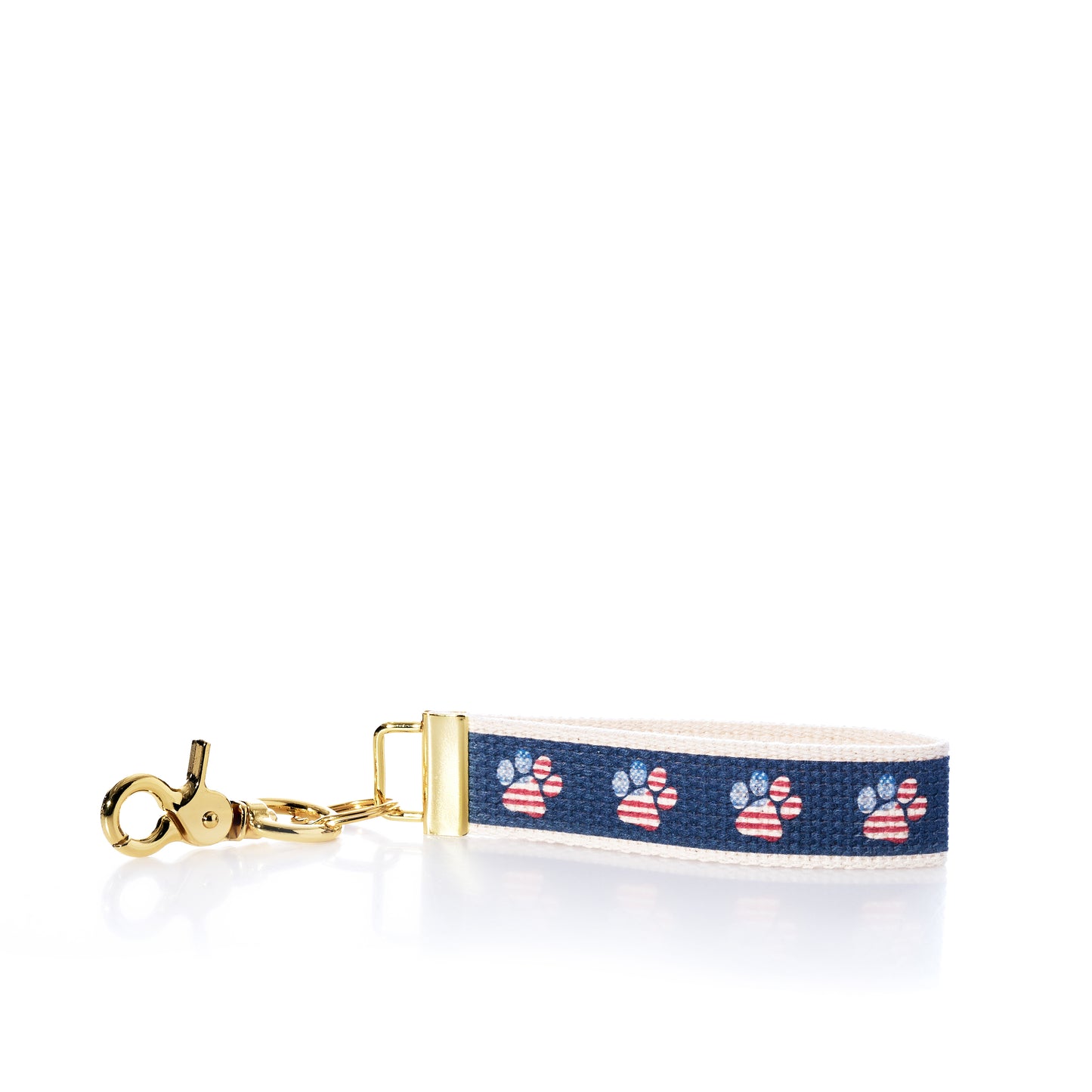 The Rustic Marlin Keychain - American Flag Paw by Rustic Marlin is a keychain wrist strap with a gold clasp, featuring a navy ribbon adorned with red, white, and blue paw prints on a white backing—perfect as a charming dog keychain or patriotic pawprint bag charm. Shown photographed on a white background. by Sea Bags