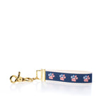 The Rustic Marlin Keychain - American Flag Paw by Rustic Marlin is a keychain wrist strap with a gold clasp, featuring a navy ribbon adorned with red, white, and blue paw prints on a white backing—perfect as a charming dog keychain or patriotic pawprint bag charm. Shown photographed on a white background. by Sea Bags