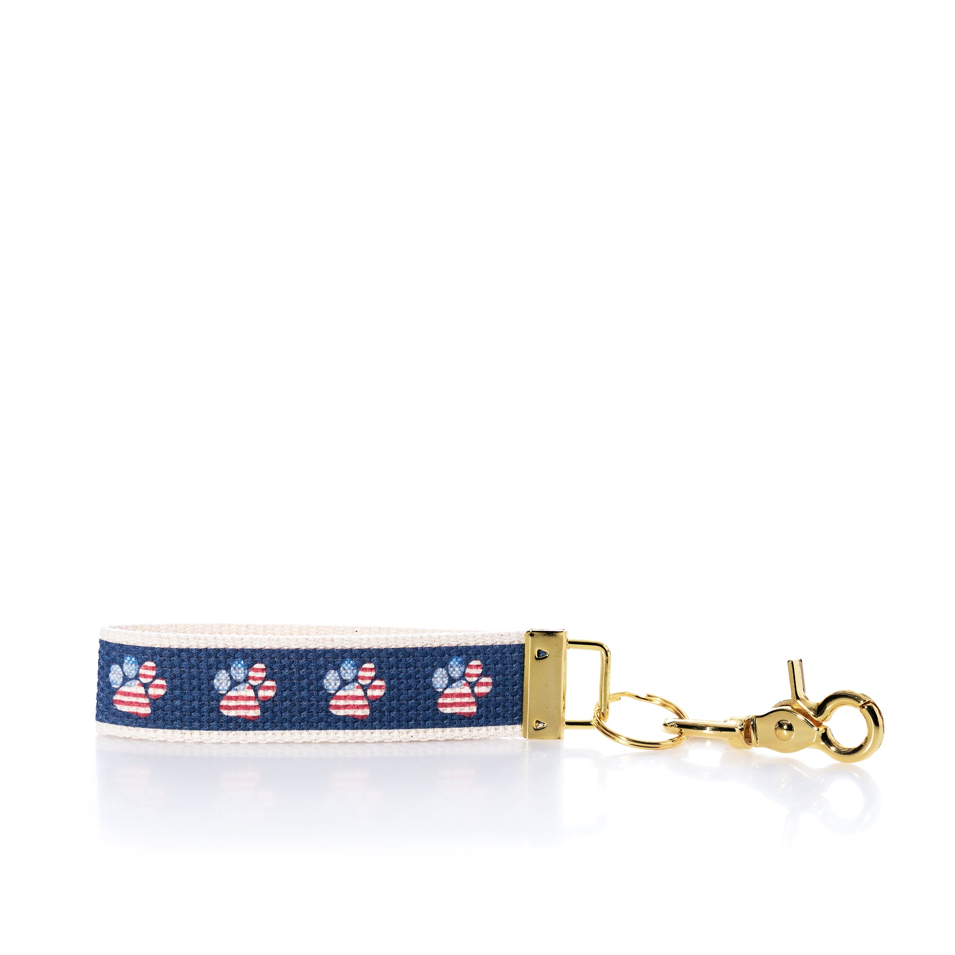 The Rustic Marlin Keychain - American Flag Paw from Rustic Marlin features a navy blue strip with American flag paw prints on a white background, complemented by a gold clasp and hardware—making it the perfect pawprint bag charm or dog keychain for any pet lover. by Sea Bags