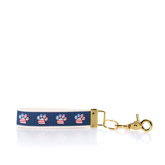 The Rustic Marlin Keychain - American Flag Paw from Rustic Marlin features a navy blue strip with American flag paw prints on a white background, complemented by a gold clasp and hardware—making it the perfect pawprint bag charm or dog keychain for any pet lover. by Sea Bags