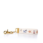 Introducing the Rustic Marlin Keychain - Summer Icons from Rustic Marlin. This keychain wrist strap showcases nautical-themed icons such as an anchor, starfish, lighthouse, and seashell printed on a white fabric band, complete with a gold clasp. The versatile design makes it perfect not just as a keyholder but also as a stylish bag charm or a unique souvenir for your collection. by Sea Bags