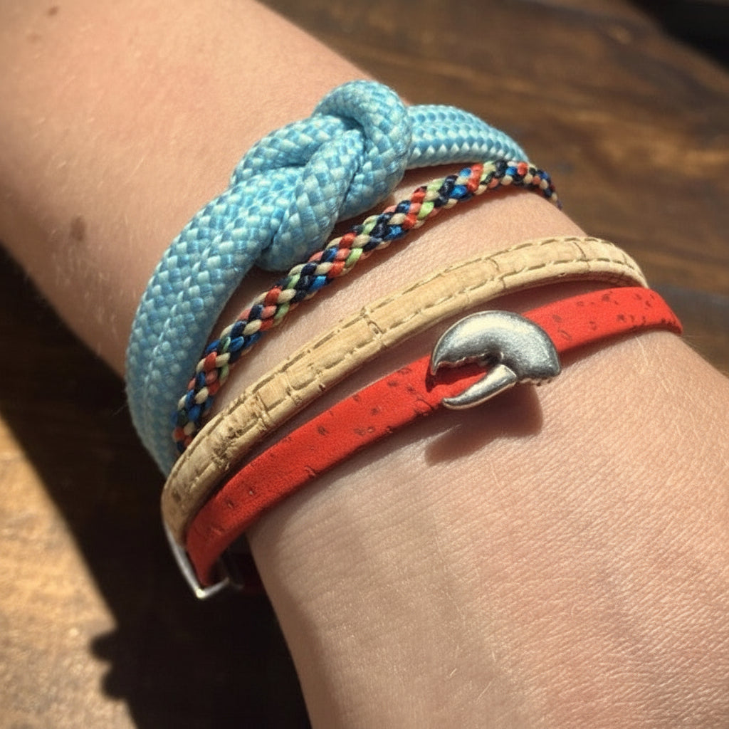A wrist adorned with four bracelets: a light blue knotted cord, a multicolored braided cord, a tan Vermont-made bracelet with a magnetic nickel-free clasp, and the Classic Cork Bracelet - Claw on Red by Elisabetta Studio featuring a silver dolphin-shaped charm. by Sea Bags
