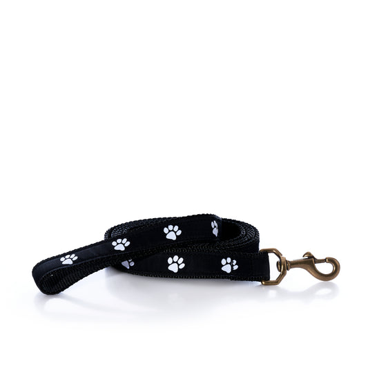 The Belted Cow Paw Print Dog Leash, featuring a black design with white paw prints and a robust metal clip, is coiled neatly on a white background.