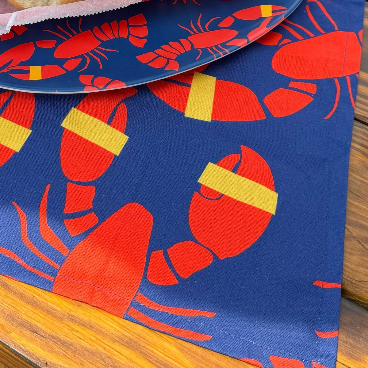 A Lobster Bake Tea Towel from Boston International, featuring a blue background with a red lobster design and yellow bands on the claws, is partly covered by a matching plate to set a charming scene reminiscent of a lobster bake on a wooden table. by Sea Bags