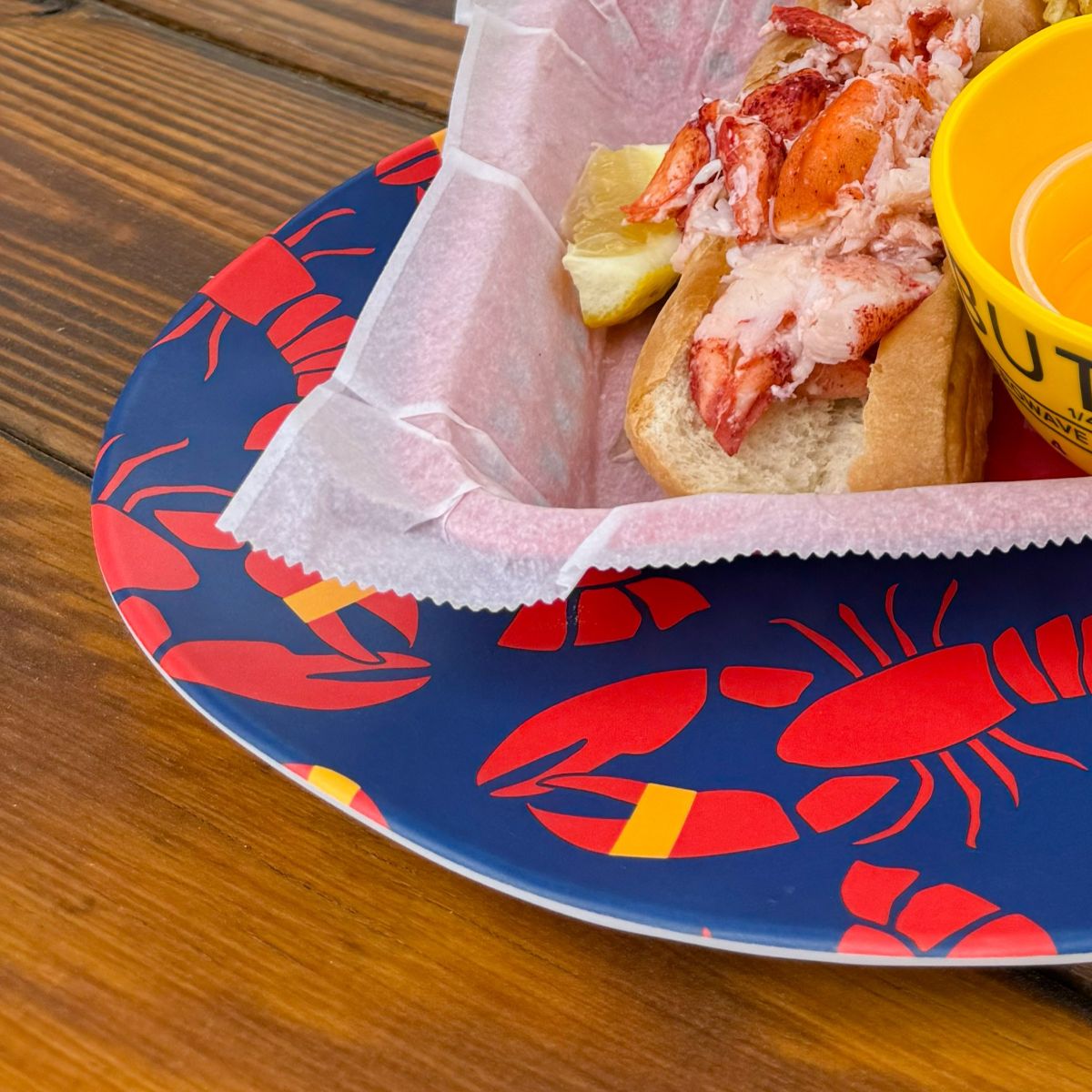 A lobster roll in a paper-lined basket sits on the Lobster Bake Serving Tray by true, featuring red lobster graphics on a blue plate, accompanied by a cup of melted butter and lemon wedges atop a wooden table.