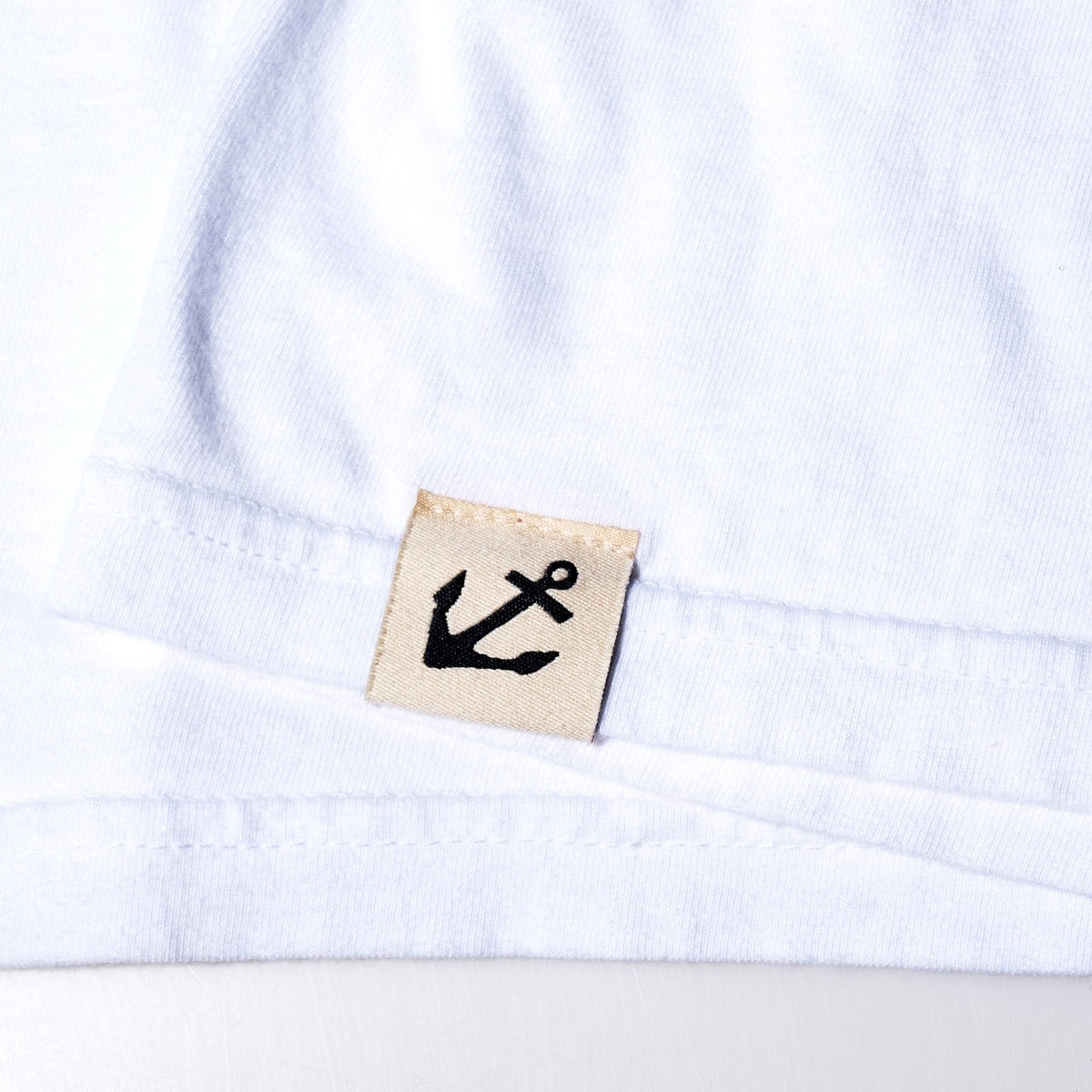 Close-up of a white fabric from the Wharf Seagull T-Shirt by 320Ink d/b/a Riptide Print Company, with a beige tag featuring a black anchor symbol, reminiscent of classic Sea Bags designs.