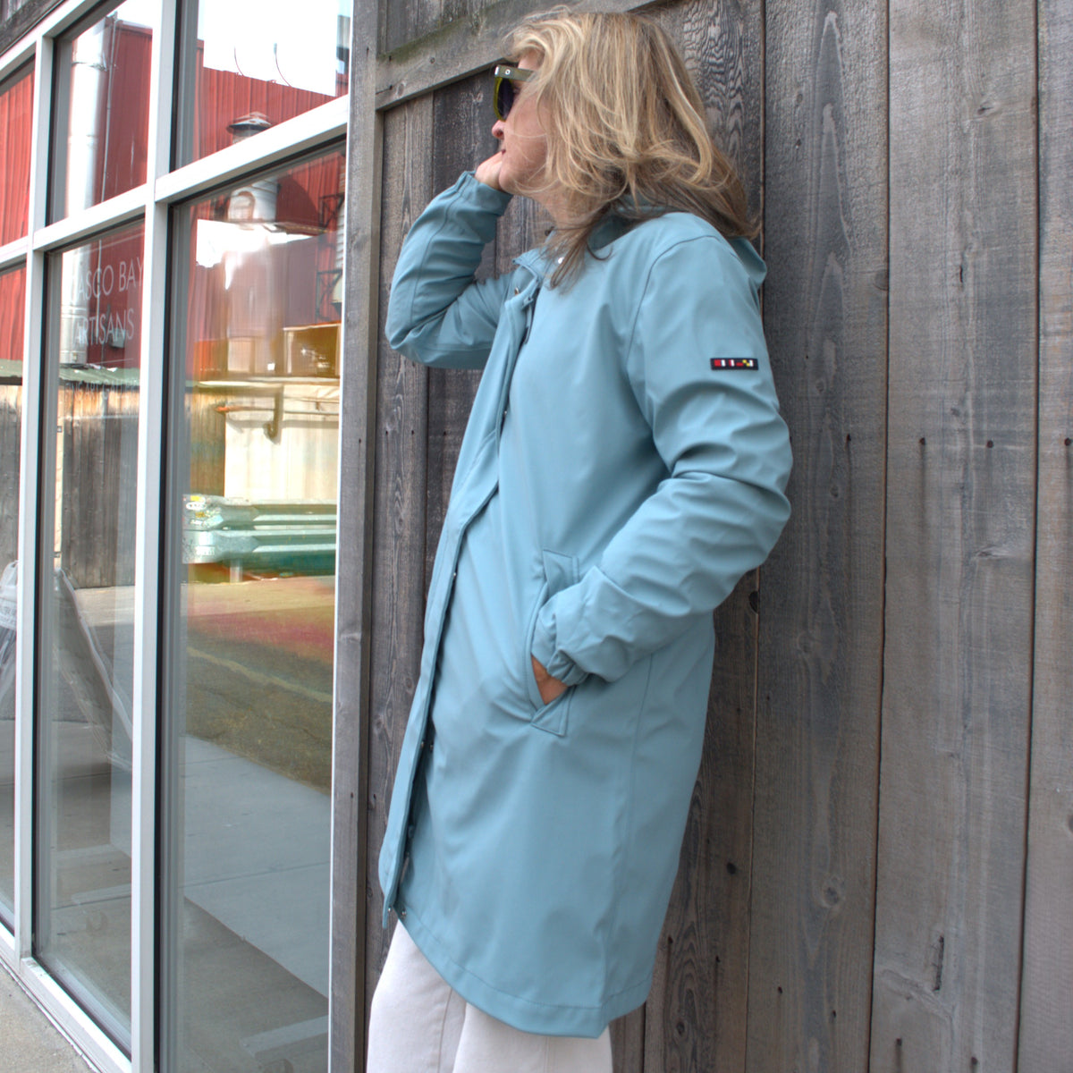 Woman in the Harborside Waterproof Raincoat - Tourmaline by Batela USA Inc stands by a wooden wall, looking away and adjusting her sunglasses.