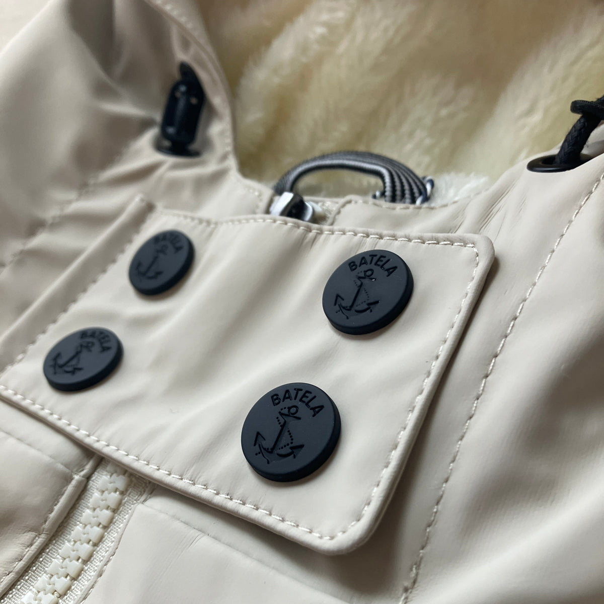 Close-up of a light beige or off-white puffer jacket with a soft cream fleece lining, featuring a front flap pocket with four dark gray BAITELA brand buttons and a silver metal zipper pull.