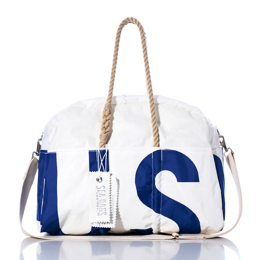 The Vintage US 52 Weekender by Sea Bags, featuring white and navy blue hues, rope handles, a white shoulder strap, and a Sea Bags tag attached, is displayed on a reflective surface. by Sea Bags