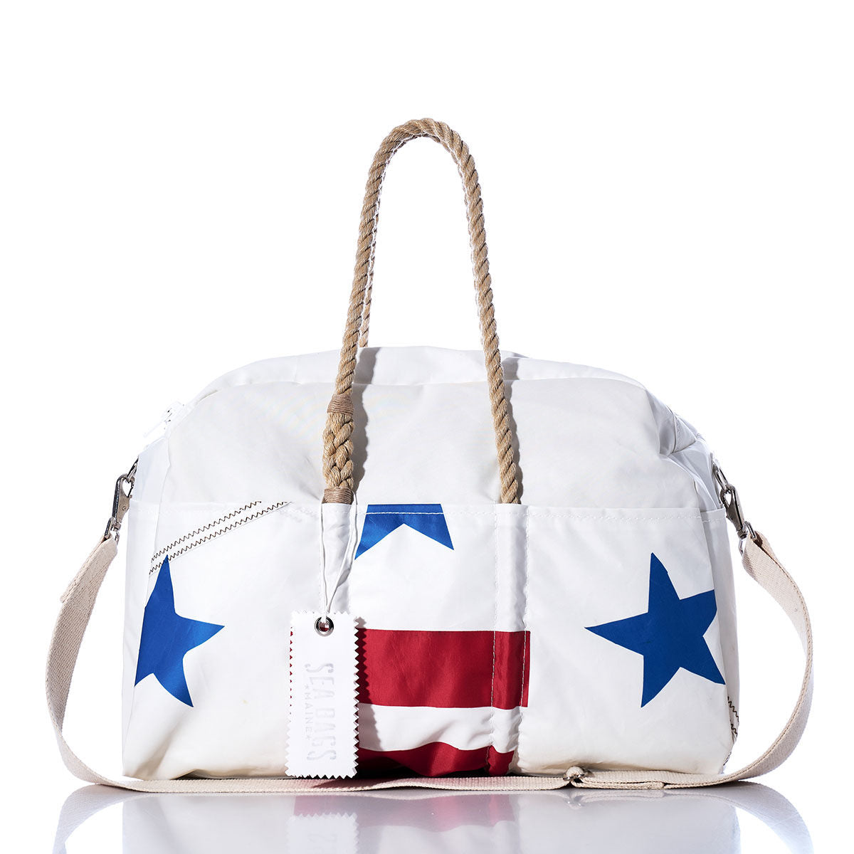 The Sea Bags Vintage Ensign Weekender features a white background adorned with blue stars and red stripes, tan rope handles, a white shoulder strap, and a white tag. Displayed on a white background, this vintage-inspired getaway bag is perfect for your next adventure. by Sea Bags