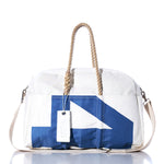 The Sea Bags Vintage 45 Weekender features a white duffel design with blue geometric print, rope handles, a beige shoulder strap, and a white tag attached to one handle. Stylishly showcased against a white background with a reflection, it’s the perfect companion for your next getaway. by Sea Bags