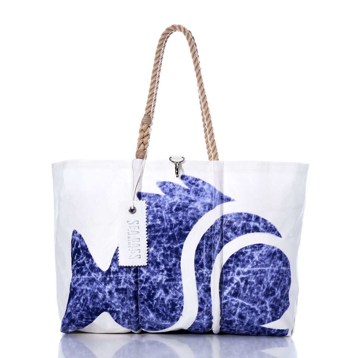 The Sea Bags Vintage Sunfish Large Tote features a white sailcloth body with a blue abstract fish motif, tan rope handles, and an attached white tag. Displayed on a reflective surface, this bag is inspired by classic Maine-made sailcloth totes. by Sea Bags