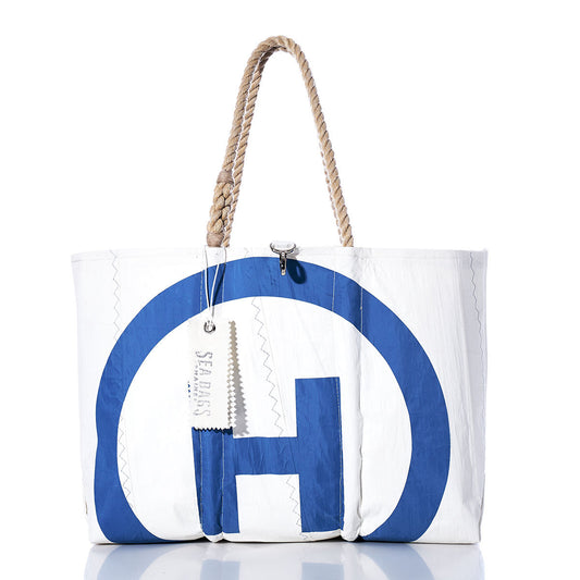 The Sea Bags Vintage Hinckley Large Tote is a white recycled sailcloth bag handcrafted in Maine, featuring a blue circular design, sturdy rope handles, and a hanging tag labeled Sea Bags. The tote stands upright on a white reflective surface. by Sea Bags