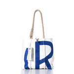 The Sea Bags Vintage Rhodes Handbag features bold blue letter print inspired by classic sail insignias, hemp rope handles, and a stitched tag. The white tote bag stands upright on a reflective surface with a white background. by Sea Bags