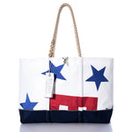 Introducing the Vintage Ensign Ogunquit Beach Tote from Sea Bags—a white tote bag crafted from recycled sail cloth, featuring blue stars, a striking red rectangle, and a navy base. Finished with rope handles and a hanging white tag with text, this stylish and eco-friendly accessory is perfect for your seaside adventures. by Sea Bags
