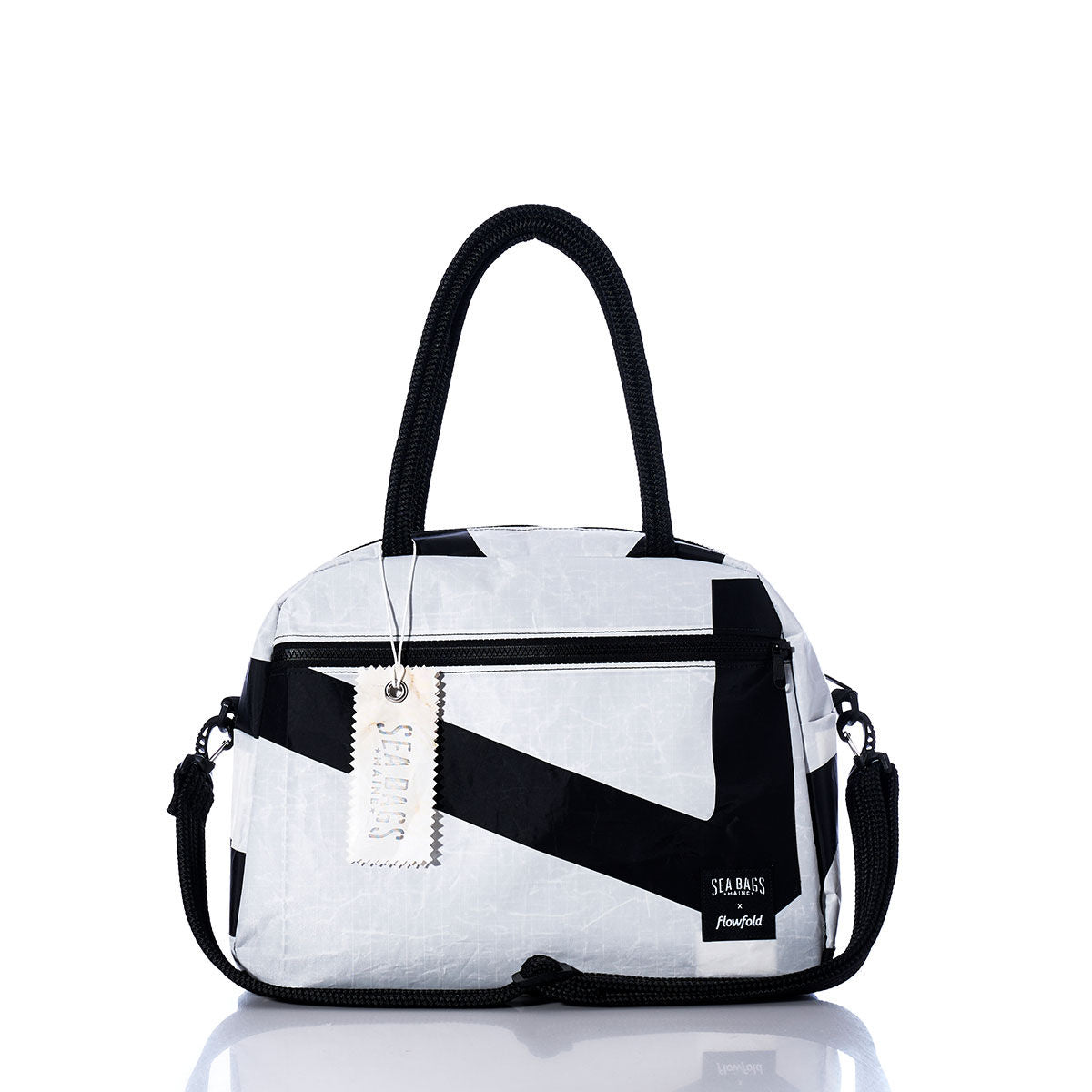 The Vintage Black and White Essential Weekender Travel Bag by Sea Bags features black handles, a front zipper pocket, an attached shoulder strap, and a branded tag reading Sea Bags. This versatile carry-on is displayed on a reflective white surface. by Sea Bags