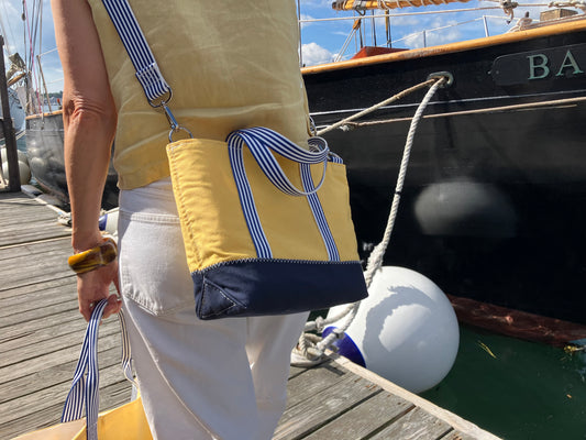 A person carrying the Sea Bags Vintage Yellow Canvas Crossbody Tote with striped straps walks along a wooden dock beside a boat, where white fenders and rope are visible. by Sea Bags