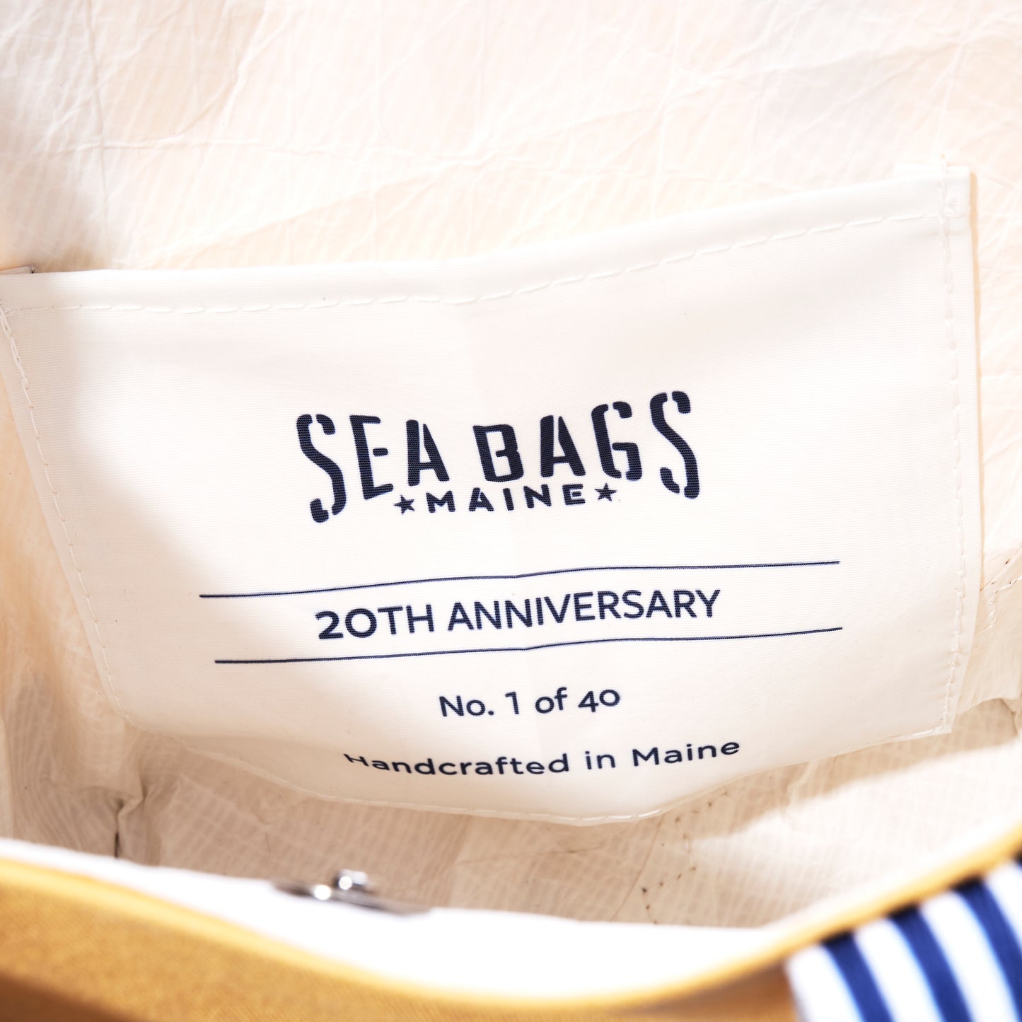 The Sea Bags Maine label inside the Vintage Yellow Canvas Crossbody Tote reads 20th Anniversary, No. 1 of 40, Handcrafted in Maine. This limited edition crossbody tote from their Heritage Collection features butter yellow canvas sail accents and blue and white striped detailing on cream-colored fabric for a distinctive yet classic design. by Sea Bags