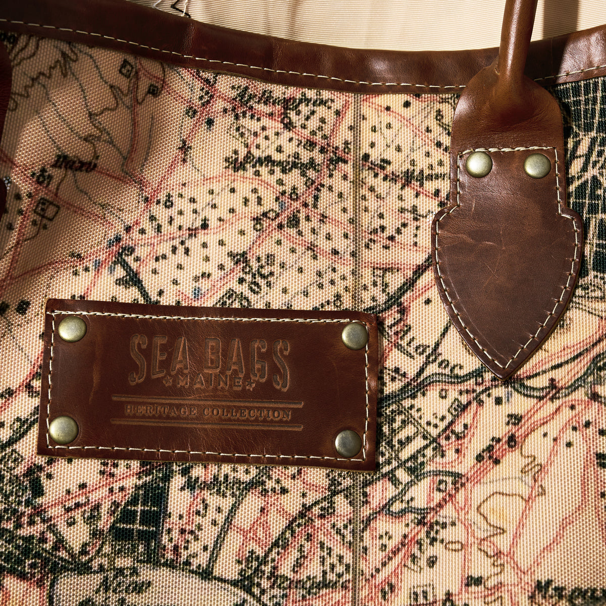 Close-up of the Sea Bags Heritage Aello Large Tote, featuring a map design, leather handle, and a leather patch embossed with “Sea Bags”—an ideal accessory for enthusiasts inspired by the classic yacht restoration traditions of the Greek sailing yacht S/Y AELLO.