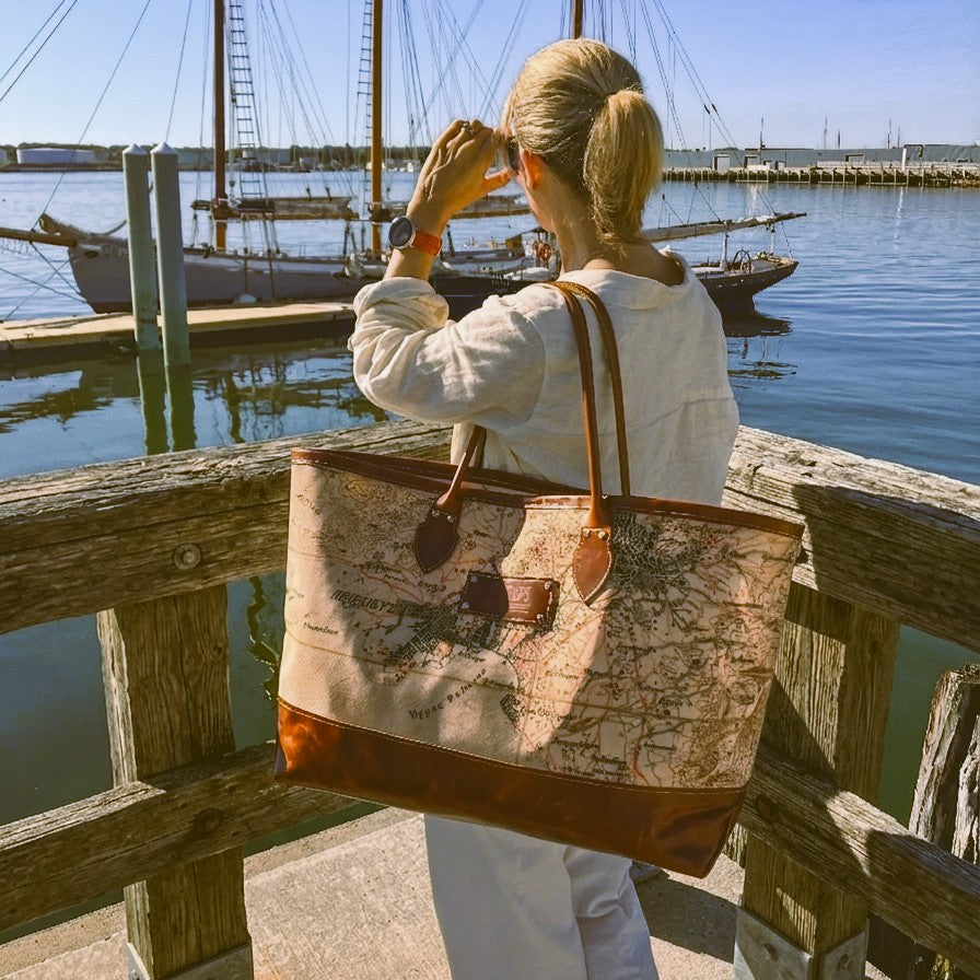 A woman carrying the Sea Bags Heritage Aello Large Tote stands on a dock, gazing at sailboats drifting on calm water beneath clear skies—including the beautifully restored S/Y AELLO, an iconic Greek sailing yacht celebrated for its classic heritage.