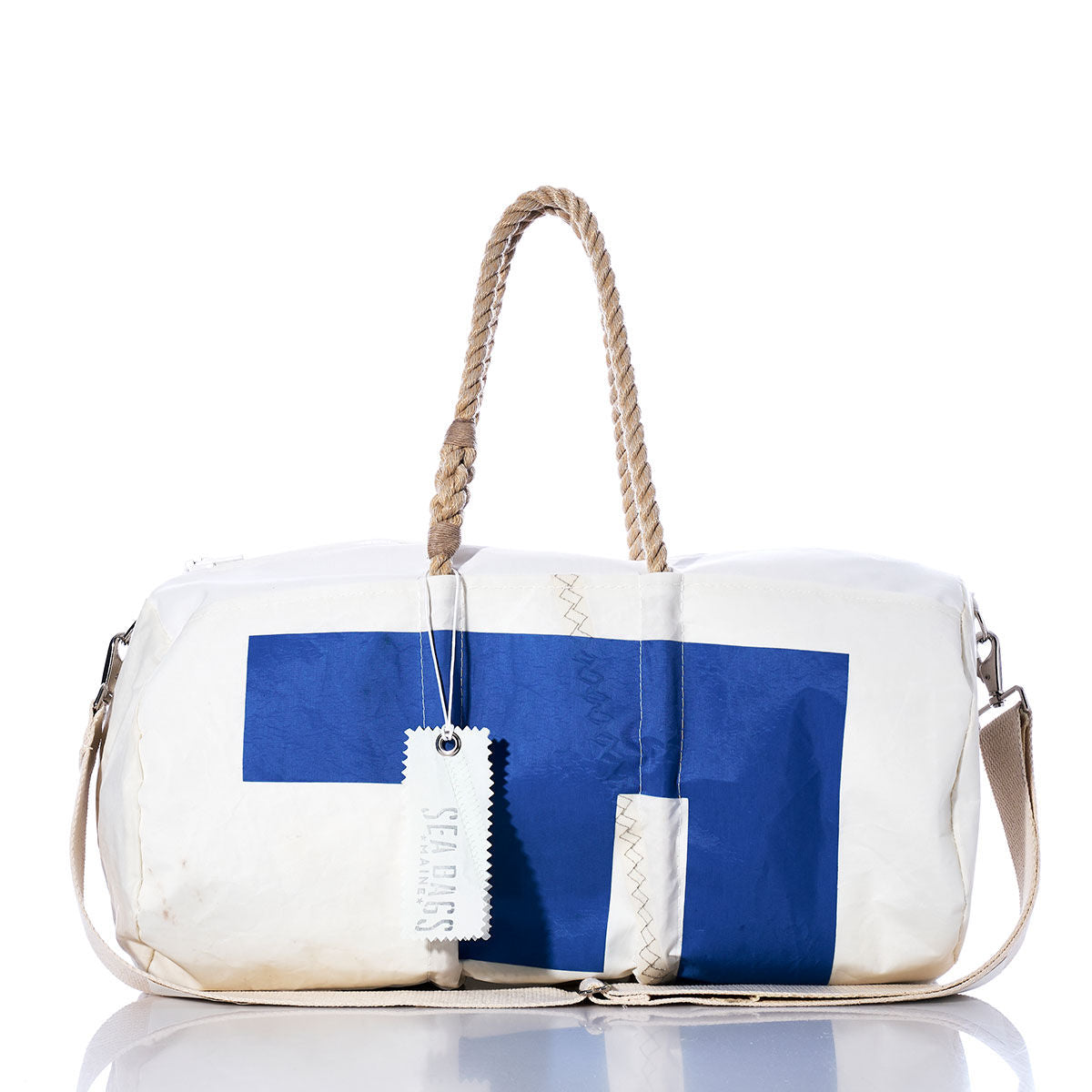 Vintage Blue F Duffel by Sea Bags, featuring a bold blue block design on white recycled sail cloth, hemp rope handles, a beige shoulder strap, and a signature white Sea Bags tag. The bag is displayed on a reflective surface against a white background. by Sea Bags