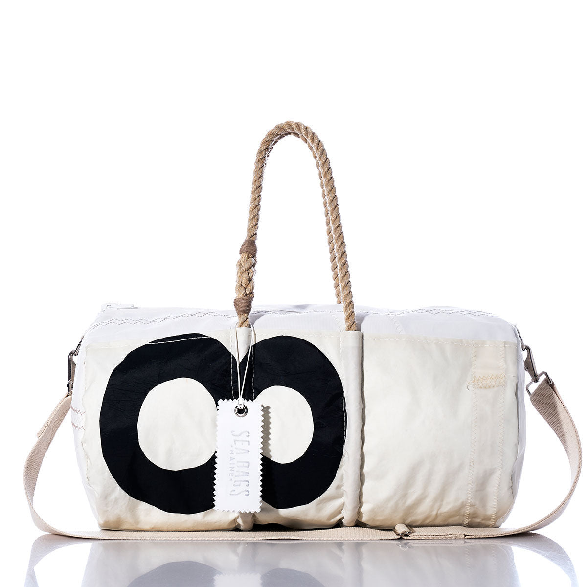 The Vintage 89 Duffel by Sea Bags is a white duffel bag featuring a bold black circular motif inspired by classic sail insignias. Made from recycled sail cloth, it comes with sturdy rope handles, a practical shoulder strap, and a signature white tag marked “SEA BAGS.” The bag is displayed against a clean white background. by Sea Bags