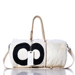 The Vintage 89 Duffel by Sea Bags is a white duffel bag featuring a bold black circular motif inspired by classic sail insignias. Made from recycled sail cloth, it comes with sturdy rope handles, a practical shoulder strap, and a signature white tag marked “SEA BAGS.” The bag is displayed against a clean white background. by Sea Bags