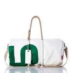 The Vintage 25 Duffel by Sea Bags is a white duffel bag made from recycled sail cloth, accented with green and red print, beige rope handles, and a shoulder strap. Sail insignias enhance its nautical appeal, while a white tag with text hangs from the handle. The vintage duffel is showcased on a reflective surface. by Sea Bags